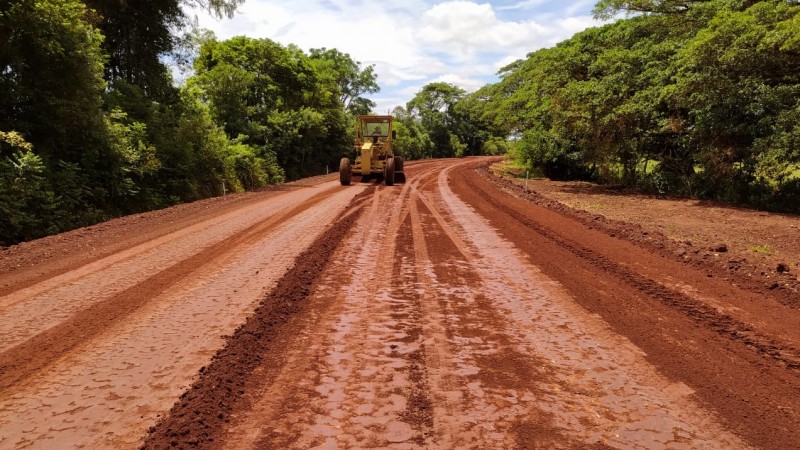 Na imagem está presente uma estrada de terra vermelha, ainda molhada pela chuva. Nos dois lados da foto aparecem várias árvores, colorindo de verde o momento. Além disso, mais ao fundo, uma patrola amarela está trabalhando.