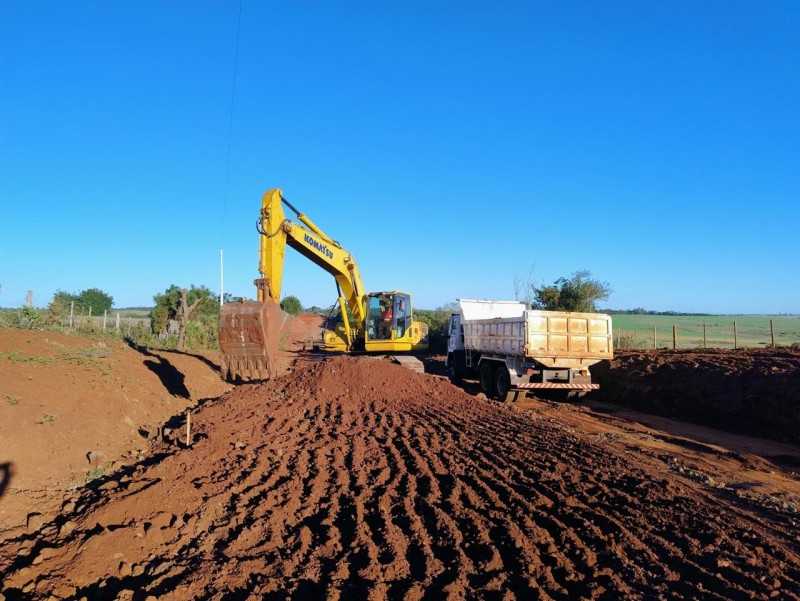 Imagem mostra obra de asfaltamento no acesso ao município de Garruchos. Na estrada estão uma retroescavadeira amarela, com um trabalhor dirigindo dentro da máquina, e um caminhão com caçamba branca estacionado.